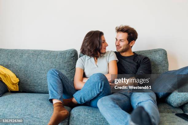 smiling couple looking at each other while sitting on sofa - coppia giovane foto e immagini stock