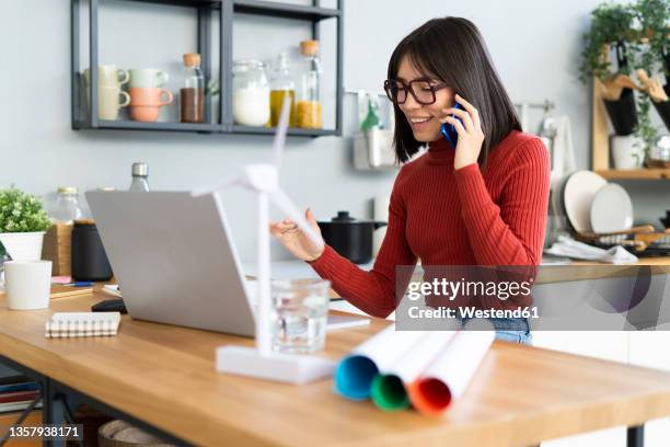 smiling freelancer with laptop talking on mobile phone at home office - green economy stockfoto's en -beelden