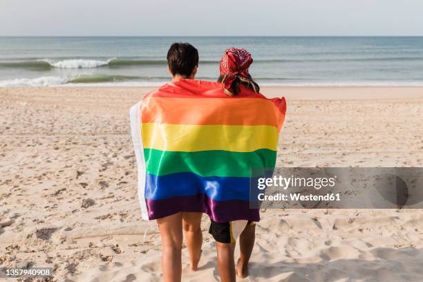 lesbian women with rainbow flag walking towards shore at beach - regenbogenfahne stock-fotos und bilder