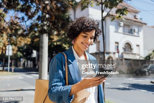 A Delighted African-American Woman Texting On Her Smartphone While Walking Through The City A Delighted African-American Woman Texting On Her Smartphone While Walking Through The City