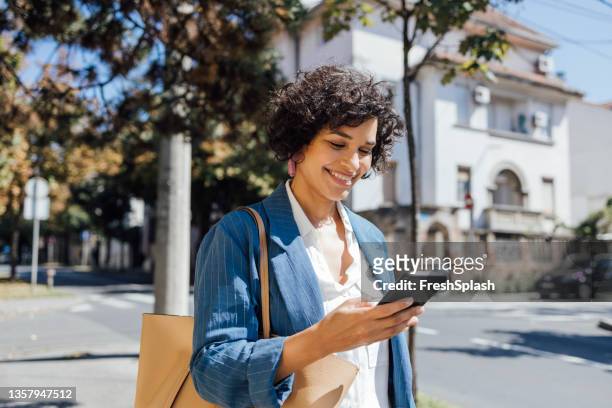 a delighted african-american woman texting on her smartphone while walking through the city - eastern european woman stock pictures, royalty-free photos & images