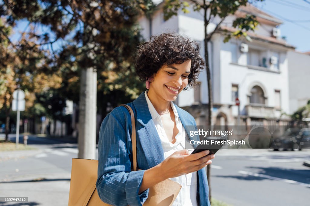 A Delighted African-American Woman Texting On Her Smartphone While Walking Through The City