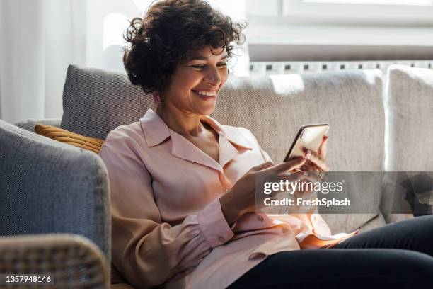 una mujer de raza mixta sentada en su sofá, viendo su podcast favorito en su teléfono inteligente y disfrutándolo - mujeres de mediana edad fotografías e imágenes de stock