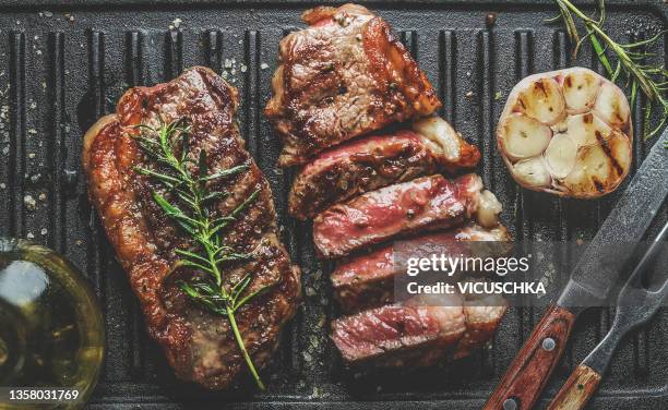 close up of sliced roasted medium rare barbecue steak with rosemary, roasted garlic and cutlery on rustic iron grill - metal grate fotografías e imágenes de stock