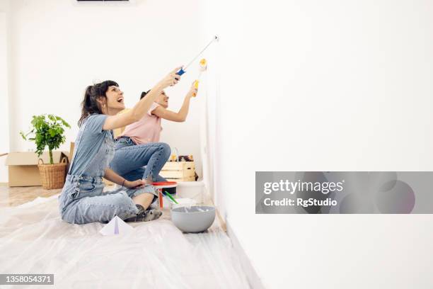 happy twin sisters painting walls while moving into their new apartment - schilderen stockfoto's en -beelden