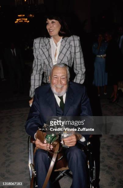 American actress Anjelica Huston pushing her father, film director John Huston , who holds an oxygen cylinder and breathing apparatus as he sits in a...