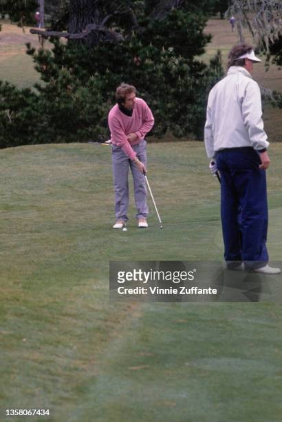 American singer, songwriter and actor Huey Lewis, wearing a pink sweater and grey trousers, lines up his putt from the edge of the green during the...