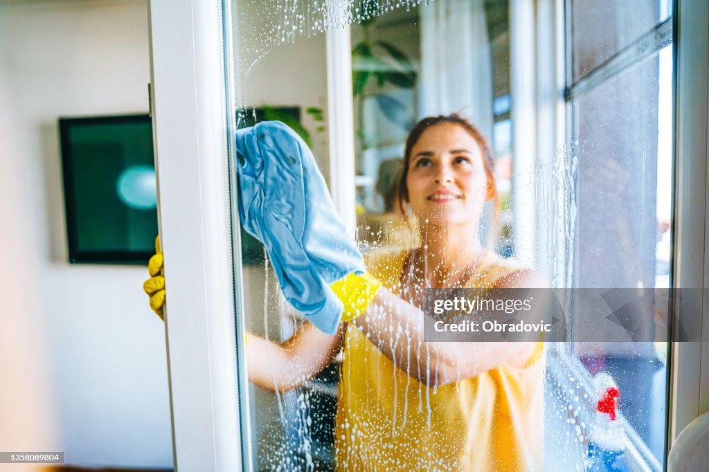 Schöne lächelnde junge Frau putzt und wischt Fenster mit Sprühflasche und Rag Stock Foto