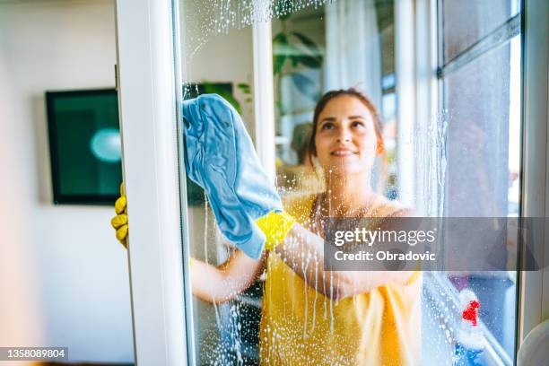 schöne lächelnde junge frau putzt und wischt fenster mit sprühflasche und rag stock foto - sterile stock-fotos und bilder