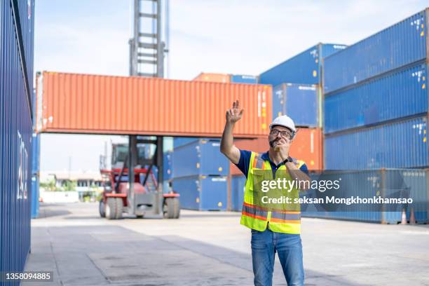 caucasian foreman man is using walkie talkie to command his workers with crane lifting container in background in container depot terminal. - box lifting technique stock-fotos und bilder
