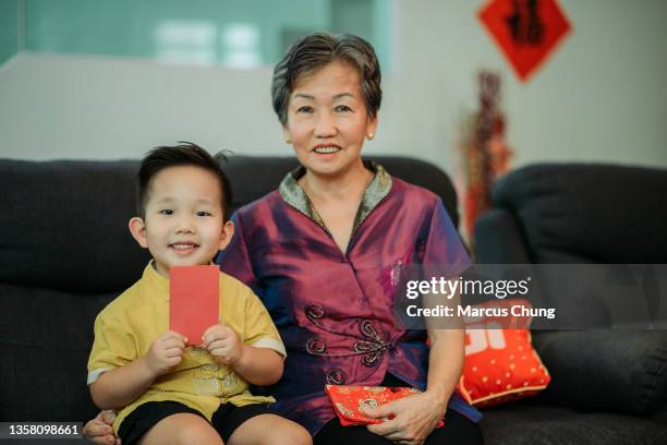 nonna e nipote cinesi asiatici sorridenti che tengono la busta rossa seduti sul divano nel soggiorno durante il capodanno cinese - abito tradizionale cinese foto e immagini stock