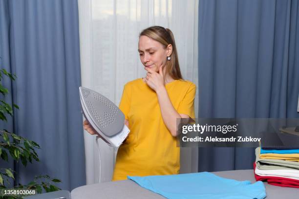 lots of bright, clean, ironed clothes folded on the ironing board. a sad, exhausted, tired woman, housewife, cleaner or housekeeper looks at a broken or faulty iron. the concept of homework, lack of time for household chores. women's duties, home life. - iron appliance stockfoto's en -beelden