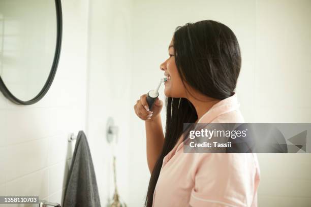 shot of a young woman brushing her teeth with an electric toothbrush at home - electric toothbrush stockfoto's en -beelden