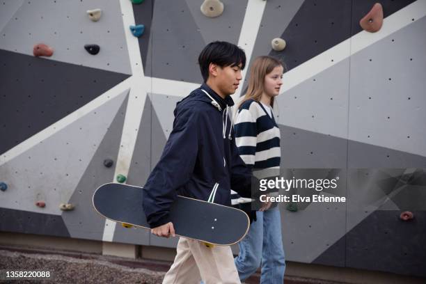 teenage girl and boy carrying a skateboard and walking past a climbing wall in a city - tcs stock pictures, royalty-free photos & images