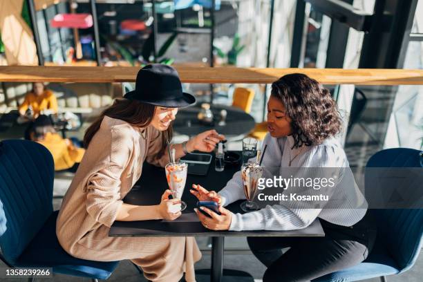 two best friends sitting in a coffee shop looking at a smart phone and smiling. - blended coffee drink stock pictures, royalty-free photos & images