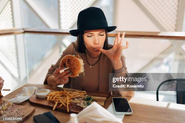 a young woman with a hat on her head sits alone at a table in a restaurant and enjoys unhealthy food. - woman eating burger stockfoto's en -beelden