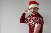 vaccine strong for Christmas. Hyped young man poses with santa claus hat and red sweater with arm bandage over silver grey background