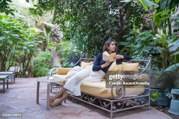 young woman relaxes on couch with book in garden - saudi relaxing stock pictures, royalty-free photos & images