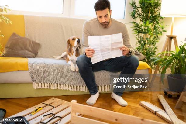 con un cane beagle accanto a lui, confuso uomo caucasico, leggendo istruzioni manuali, mentre assemblava mobili - manuale foto e immagini stock