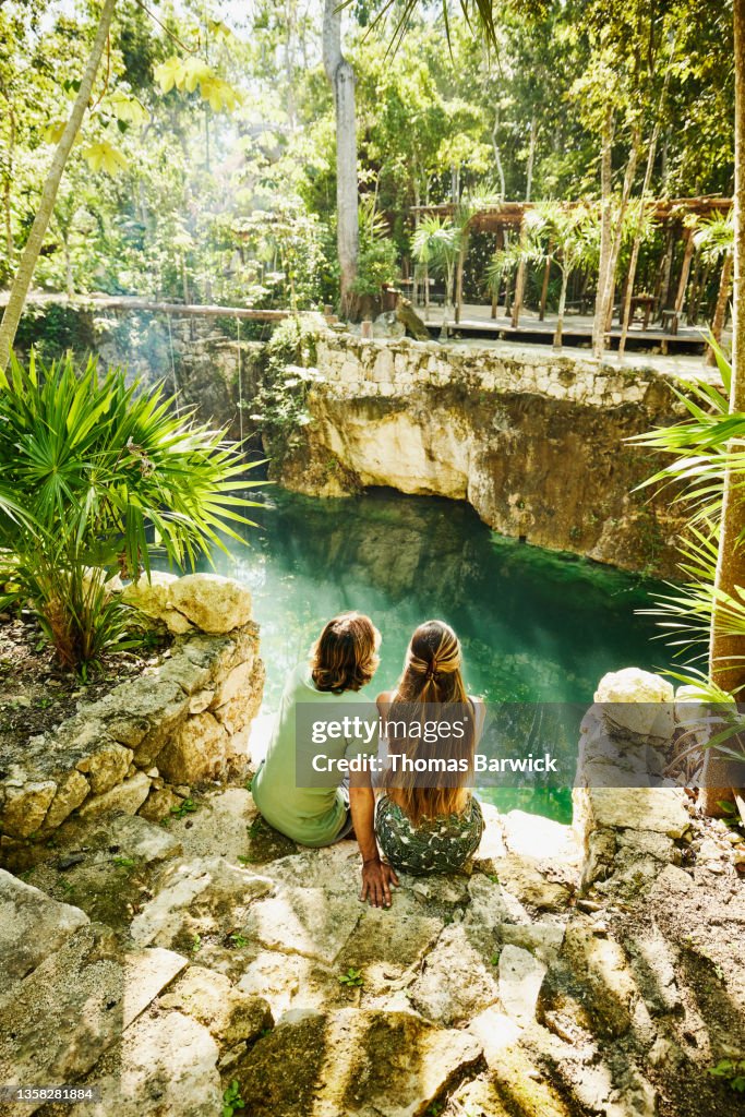 Wide shot rear view of couple relaxing at edge of cenote at eco resort in jungle