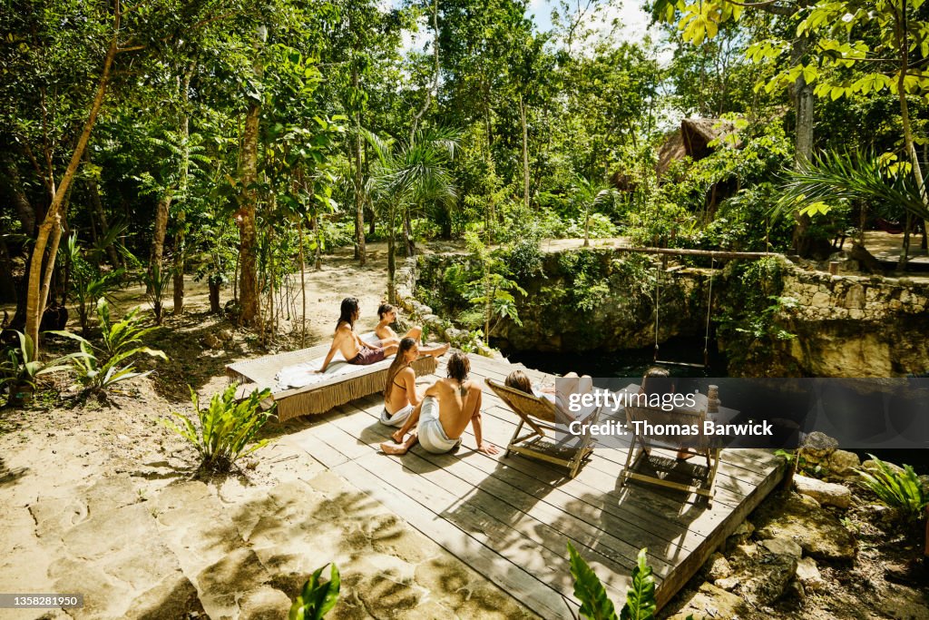 Wide shot of friends relaxing in sun on platform above cenote at eco resort in jungle