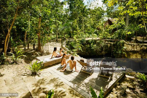 Wide shot of friends relaxing in sun on platform above cenote at eco resort in jungle