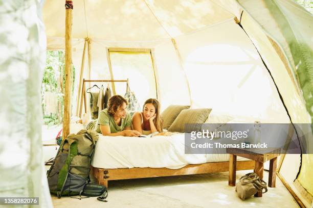 Wide shot of couple reading on bed in tent while staying at eco resort in jungle