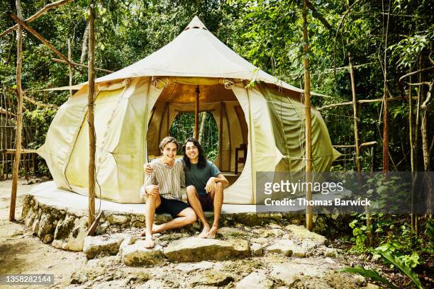 wide shot portrait of smiling gay couple sitting in front of tent while glamping at eco resort in jungle - turismo ecológico fotografías e imágenes de stock