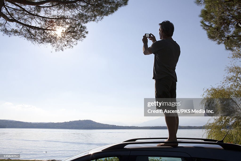 Man stands on car roof above lake, takes picture