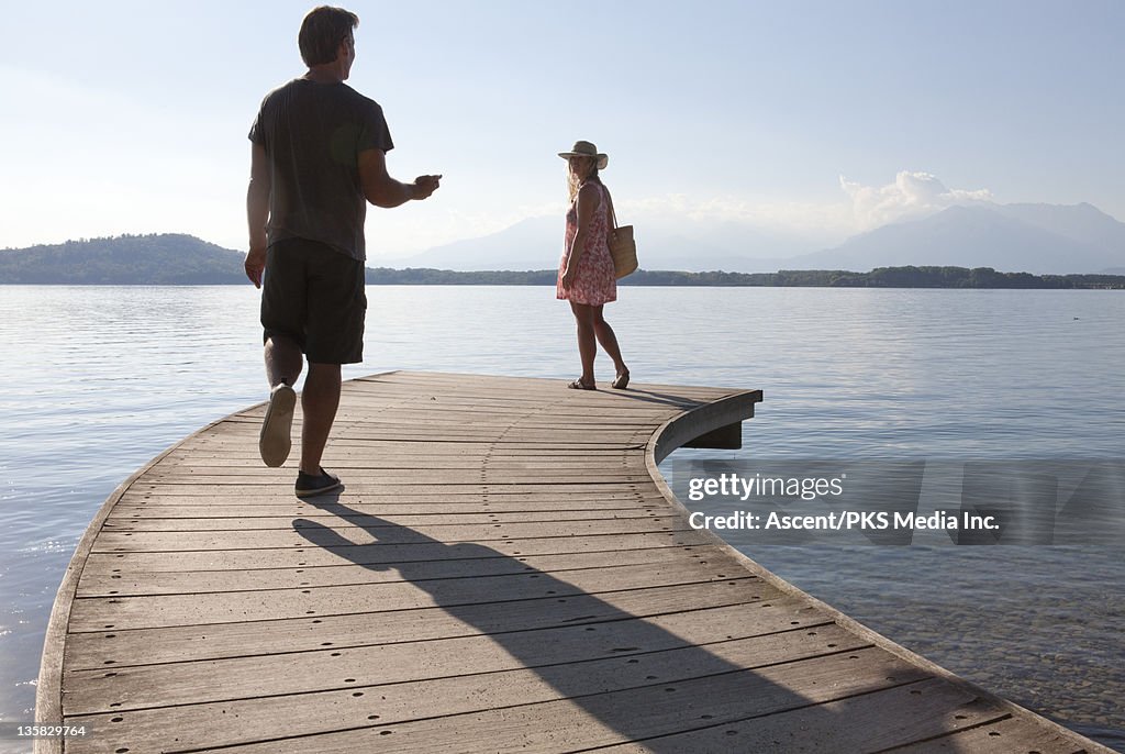 Man walks toward woman on lake wharf, holds cell