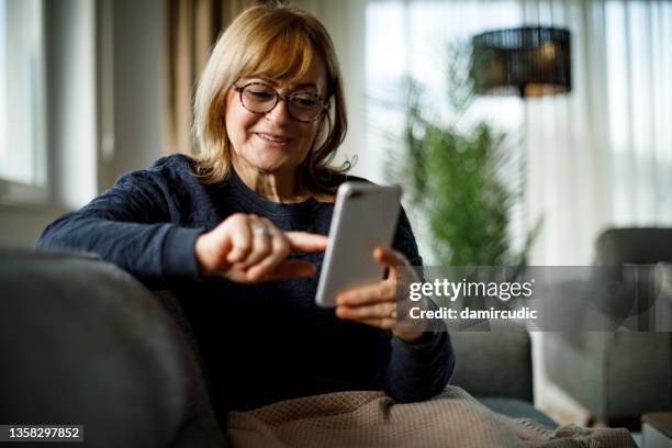 mujer madura sonriente usando el teléfono móvil en casa - choose fotografías e imágenes de stock
