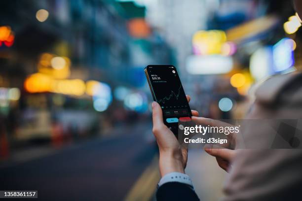 over the shoulder view of young asian woman managing financial stock trading with online banking account on smartphone on the go, city scene with illuminated and multi-coloured neon signboards in background. business on the go - mobile money stock-fotos und bilder