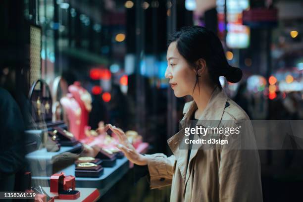 smiling young asian woman standing outside a boutique looking at gorgeous jewellery on the shop window in the city at night - window shopping stock-fotos und bilder