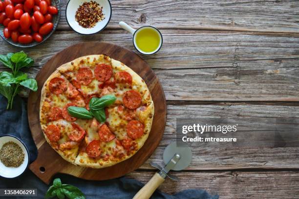 image of sliced pepperoni pizza on round wooden chopping board, melted golden buffalo mozzarella cheese, rich tomato marinara sauce and fresh basil leaf topping, blue tea towel, pizza wheel, wood grain background, copy space - round wooden chopping board stock pictures, royalty-free photos & images