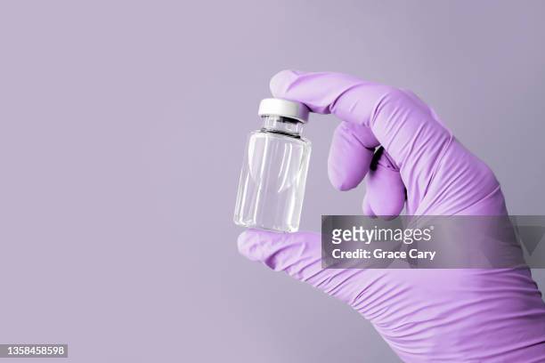 hand wearing purple glove holds vial filled with clear liquid against purple background - studio shot stockfoto's en -beelden