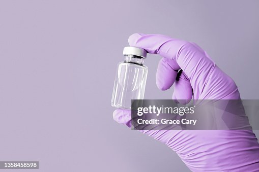 Hand Wearing Purple Glove Holds Vial Filled With Clear Liquid Against Purple Background Hand Wearing Purple Glove Holds Vial Filled With Clear Liquid Against Purple Background