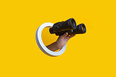 Woman's hand holding binoculars in a hole on a yellow background.