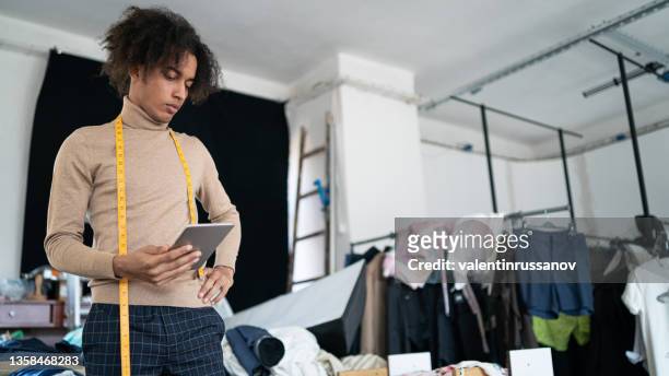 serious young african fashion designer keeping hand on his hip and holding a tablet in the other hand, looking at  fabrics and patterns in his workshop - clothing design studio stock pictures, royalty-free photos & images