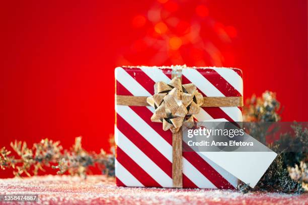 striped white-red gift box with white tag and gold bow near christmas tree branches on red table with artificial snow against red wall with defocused garland. new year coming concept. front view and close-up - christmas tag stockfoto's en -beelden