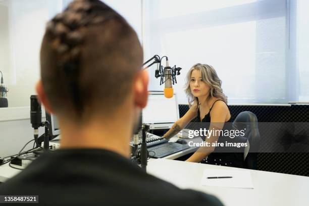 front view of beautiful presenter wearing black clothing sitting in front of the microphone looking away while interview male artist during a radio show - radio station fotografías e imágenes de stock