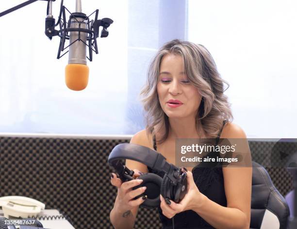 front view of beautiful blonde female host sitting in front of a microphone of a broadcast room of a radio station smiling while holding her headphones - radio host imagens e fotografias de stock