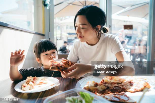 joyful young asian mother and lovely little daughter enjoying pizza lunch in a restaurant. mother is feeding daughter a slice of pizza. family enjoying bonding time and a happy meal together. family and eating out lifestyle - mother daughter brunch bildbanksfoton och bilder