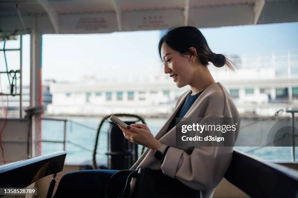 beautiful smiling young asian woman using smartphone while taking a star ferry ride in victoria harbour. lifestyle and technology - commuter ferry stock pictures, royalty-free photos & images