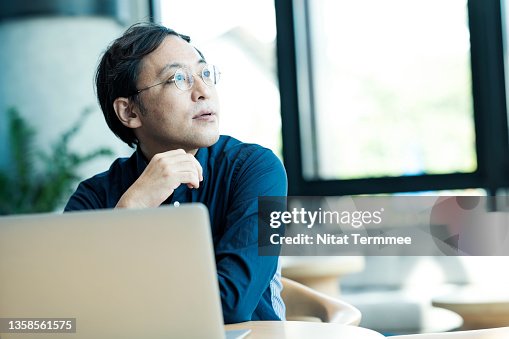 Japanese Entrepreneur using a laptop and thinking about business plan in a remote office. Business Minded Entrepreneurship. Japanese Entrepreneur using a laptop and thinking about business plan in a remote office. Business Minded Entrepreneurship.