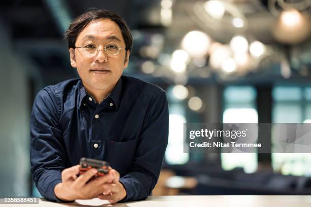 portrait of a japanese entrepreneur standing and holding a mobile phone in a modern office. corporate business, business entrepreneurship. - asian businessman phone stockfoto's en -beelden