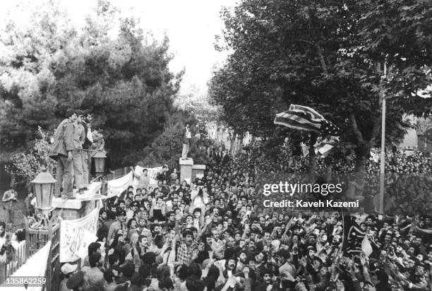 Standing on the walls of the compound, Iranian students following the Imam Khomeini line throw a US flag into the crowds gathering outside the...