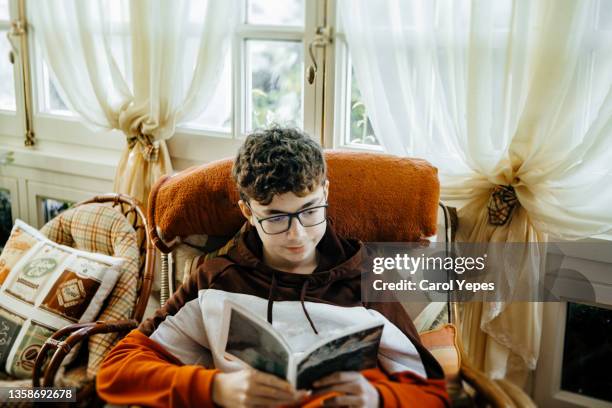 hispanic boy in eyeglasses reading a book at living room - one teenage boy only stock pictures, royalty-free photos & images