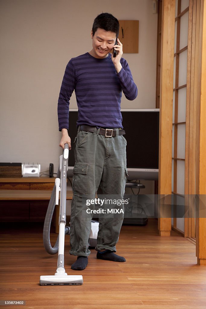 Young man vacuuming floor and holding cellular phone
