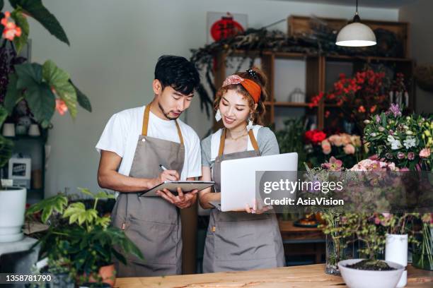 joven pareja asiática, propietarios de una pequeña empresa de floristería, usando computadora portátil y trabajando juntos en la tienda contra flores y plantas. discutir asuntos de negocios. start-up business, asociación empresarial y trabajo en equip - asia lady selling flower fotografías e imágenes de stock