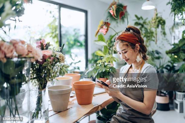 young asian female florist, owner of small business flower shop, using smartphone at shop counter against flowers and plants. checking stocks, taking customers orders, selling products online. daily routine of running a small business with technology - asia lady selling flower stock pictures, royalty-free photos & images